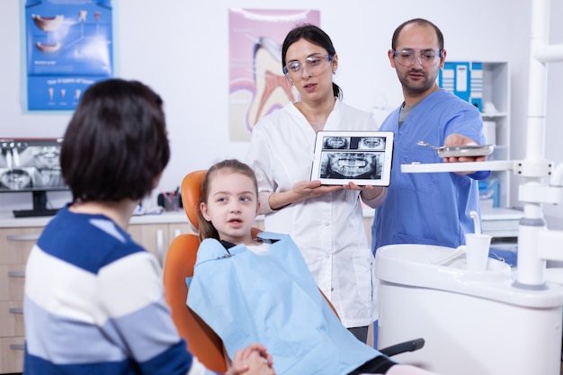 Parent avec une petite fille dans un cabinet dentaire écoutant un médecin dentiste parlant du traitement de la carie. Stomatologue expliquant le diagnostic des dents à la mère de l'enfant dans une clinique de santé tenant une radiographie.