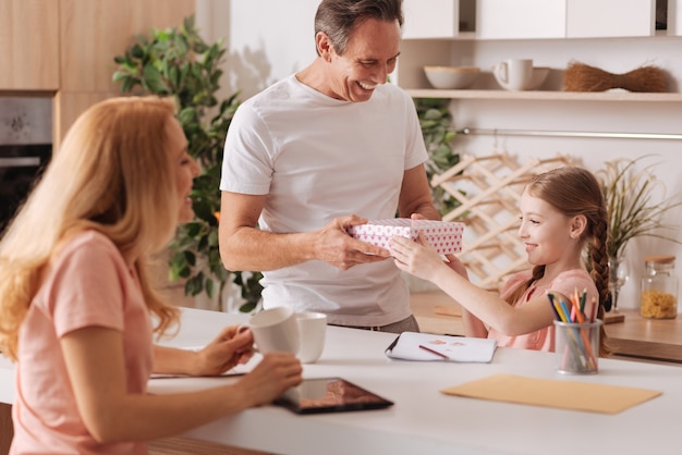 Les parents aimants ravis d'âge mûr profitant de vacances à la maison et se reposant tout en donnant une boîte-cadeau à la fille