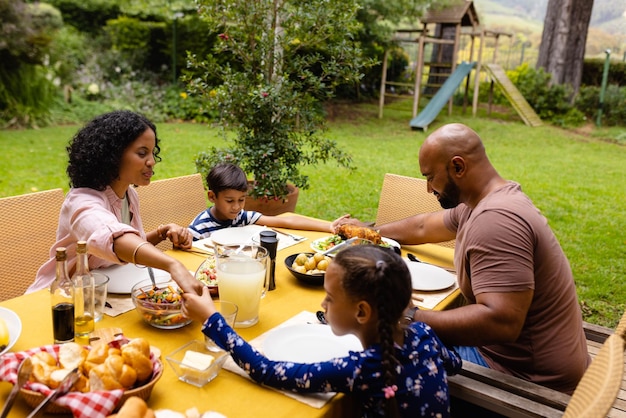 Des parents biraciaux heureux, un fils et une fille priant avant le repas, se tenant par la main à table dans le jardin.