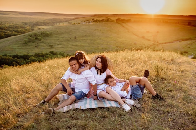 Les parents et deux enfants sont assis sur un tapis sur une colline au coucher du soleil en été