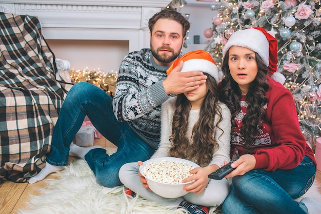 Les parents effrayés ont l'air tout droit. Le père couvre les yeux de sa fille avec la main. Ils regardent ensemble. Fille tient un bol de pop-corn. Elle est assise entre les parents.