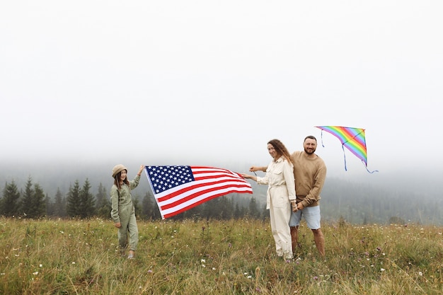 Les parents et les enfants avec le drapeau américain jouent avec un cerf-volant coloré. mère, père et petite fille célèbrent ensemble le 4 juillet à l'extérieur par temps brumeux. Concept de la fête de l'indépendance des Etats-Unis.