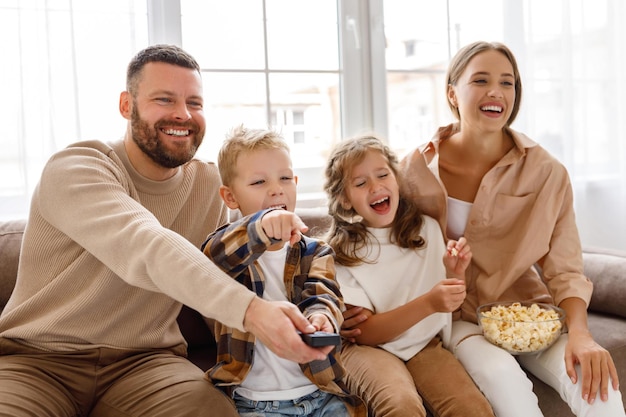 Des parents et des enfants de famille ravis mangeant du pop-corn et riant assis sur un canapé et regardant un film amusant à la maison