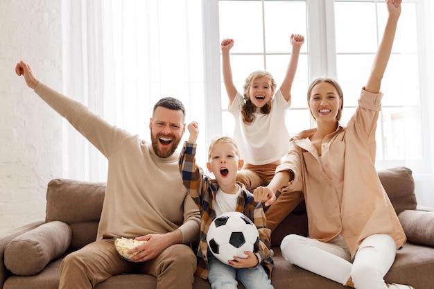 Des parents et des enfants ravis avec du pop-corn et une balle levant les bras et criant tout en regardant un match de football et en célébrant le but à la maison