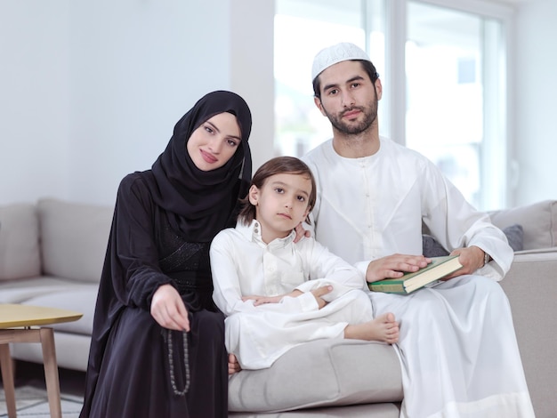 Parents de famille musulmans traditionnels avec enfants lisant le Coran et priant ensemble sur le canapé avant le dîner de l'iftar lors d'une fête du ramadan à la maison