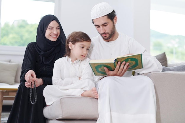 Parents de famille musulmans traditionnels avec enfants lisant le Coran et priant ensemble sur le canapé avant le dîner de l'iftar lors d'une fête du ramadan à la maison