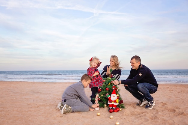 Parents Avec Fils Et Petite Fille Décorant L'arbre De Noël Sur La Plage Au Bord De La Mer.