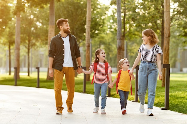 Parents joyeux et enfants ravis avec des sacs à dos tenant la main et souriant tout en marchant à l'école par une journée ensoleillée dans le parc