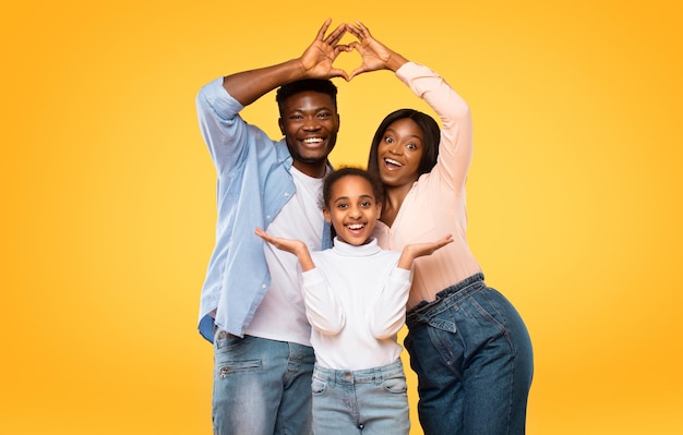 Photo parents noirs excités montrant la forme du cœur avec les doigts faisant un toit symbolique sous la tête de leur fille