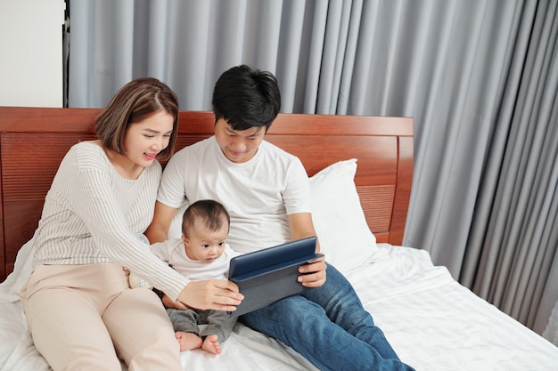 Parents et petit enfant regardant un dessin animé intéressant sur une tablette en se reposant sur le lit le soir