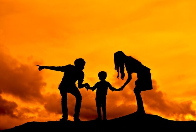 Photo des parents en silhouette tenant la main de leur fils sur la montagne contre un ciel nuageux orange au coucher du soleil