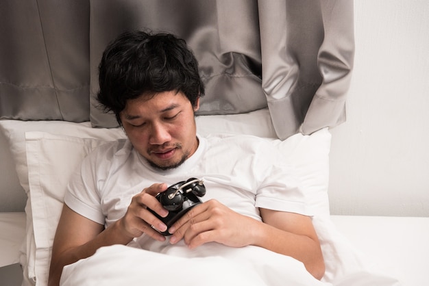 Paresse de l&#39;homme asiatique qui dort encore sur le lit dans la chambre du matin avec réveil noir