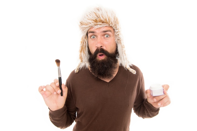 Parfois, la mode devient bizarre. Et si ajouter du maquillage. Un styliste de mode barbu porte un chapeau à fourrure tenir une brosse pour se maquiller. Soins du visage. Dernières tendances de la mode. Mec freak excentrique isolé sur blanc.