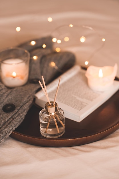 Parfum d'ambiance liquide dans une bouteille en verre avec des bâtons, un livre en papier ouvert, des bougies parfumées allumées et du textile en laine tricoté au lit en gros plan. Saison des vacances d'hiver. Ambiance chaleureuse. Aromathérapie.