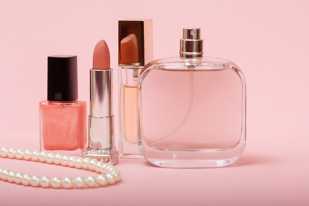 Parfum femme, perles, flacon avec vernis à ongles et rouge à lèvres sur fond rose. Cosmétiques et parfums pour femmes.