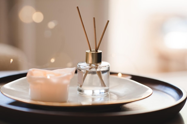 Parfum parfumé liquide maison dans une bouteille en verre avec des bâtons de bambou sur une assiette avec une bougie allumée dans la chambre en gros plan Ambiance chaleureuse