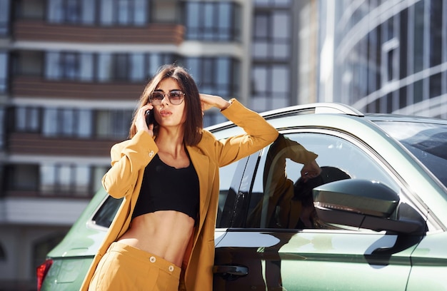 Parle au téléphone Jeune femme à la mode en manteau de couleur bordeaux pendant la journée avec sa voiture