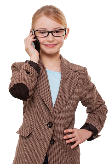 Parler d'affaires. Joyeuse petite fille en tenue de soirée parlant au téléphone portable et tenant la main sur la hanche en se tenant debout isolée sur blanc