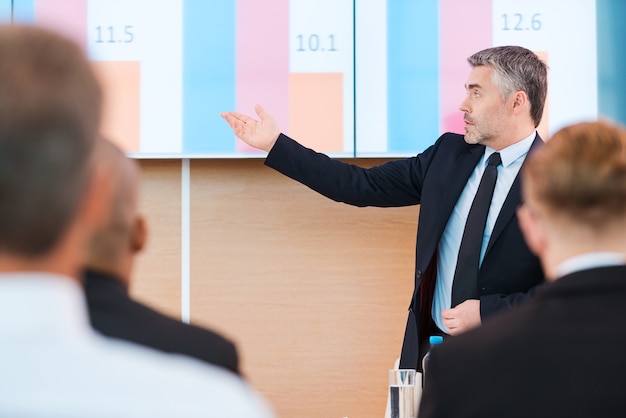 Photo parler de chiffres. homme mûr confiant en tenue de soirée pointant un écran de projection avec un graphique dessus tout en faisant une présentation dans une salle de conférence avec des personnes au premier plan