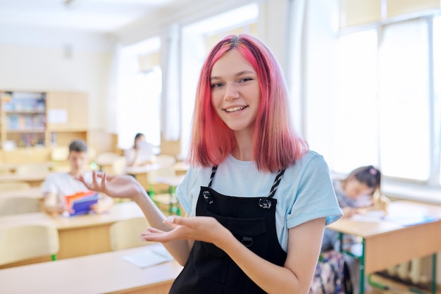 Parler en gesticulant adolescente souriante à la leçon en classe, belle fille à la mode regardant la caméra, étudiants de fond assis à des bureaux