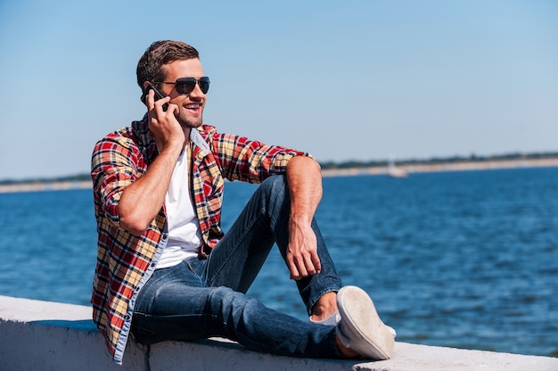 Parler sans souci. Beau jeune homme à lunettes de soleil parlant au téléphone portable et souriant alors qu'il était assis au bord de la rivière