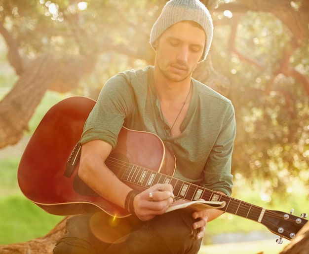 Paroles inspirées par la nature Photo d'un beau jeune homme assis dans un parc avec une guitare écrivant une chanson sur un bloc-notes