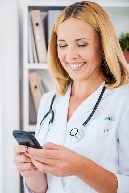 Partager de bonnes nouvelles. Joyeuse femme médecin en uniforme blanc tenant un téléphone portable et souriant