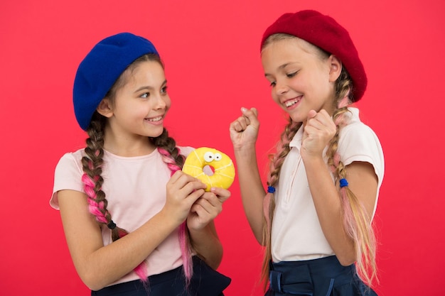 Partagez un beignet sucré. Les filles en chapeaux de béret tiennent un fond rouge de beignet glacé. Les filles enjouées sont prêtes à manger des beignets. Amitié et générosité. Concept de magasin de bonbons et de boulangerie. Les enfants sont de grands fans de beignets au four.