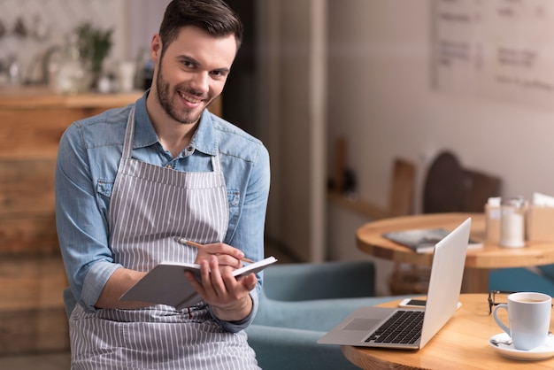 Partagez la positivité. Heureux beau jeune homme prenant des notes et souriant tout en utilisant un ordinateur portable