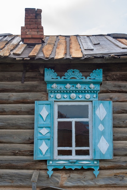 Photo partie de l'ancienne maison en rondins russe avec fenêtre et volets sculptés. sur le toit il y a une couverture pressée par des planches et une cheminée en briques. image verticale.