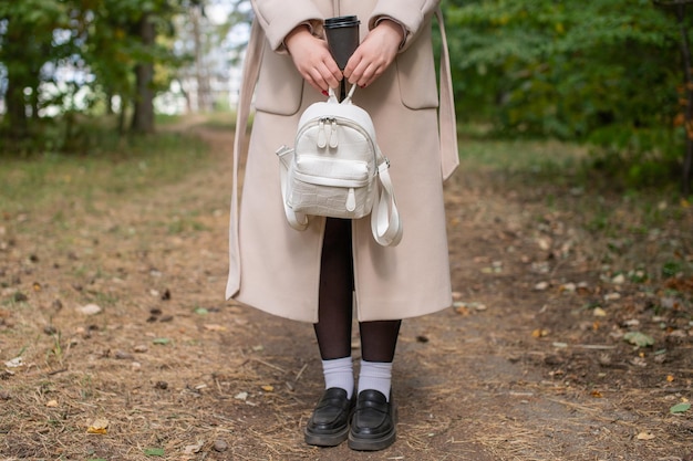 Partie du corps d'une fille dans un long manteau avec un sac à dos et une tasse de boisson dans ses mains