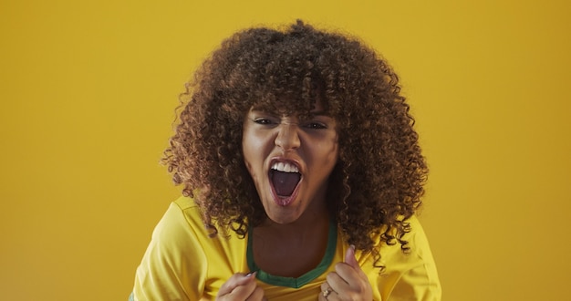 Partisan du Brésil. Fan brésilienne de cheveux bouclés célébrant le football, match de football sur fond jaune. Couleurs du Brésil.