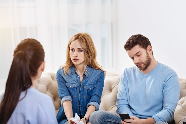 Pas disposé à coopérer. Beau jeune homme agréable assis sur le canapé et jouant sur son smartphone lors d'une visite à un psychologue avec sa femme