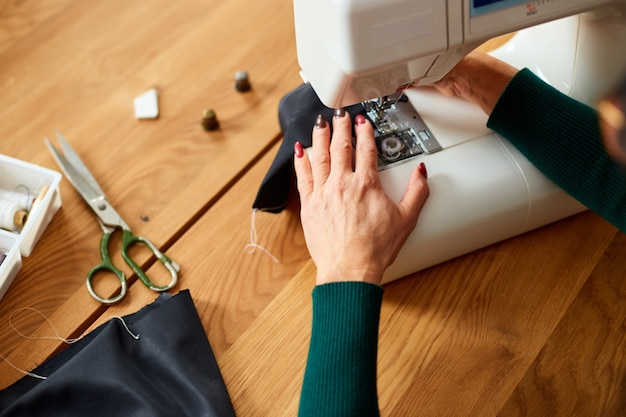 Pas à pas, une vieille de 50 ans coud des vêtements sur une machine à coudre, un tailleur mature travaillant avec la couture dans un atelier, l'industrie textile, un passe-temps, un espace de travail.