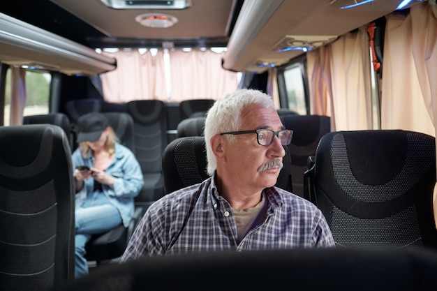 Passager senior pensif sérieux dans des verres assis dans un bus moderne et regardant par la fenêtre