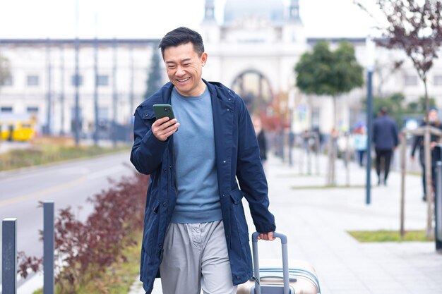 Un Passager Touristique Asiatique Arrive De L'aéroport Se Promène Dans La Ville Avec Une Valise Un Homme Sourit Et Se Réjouit Utilise Le Téléphone Et L'application Pour Réserver Un Hébergement Et Réserver Un Taxi