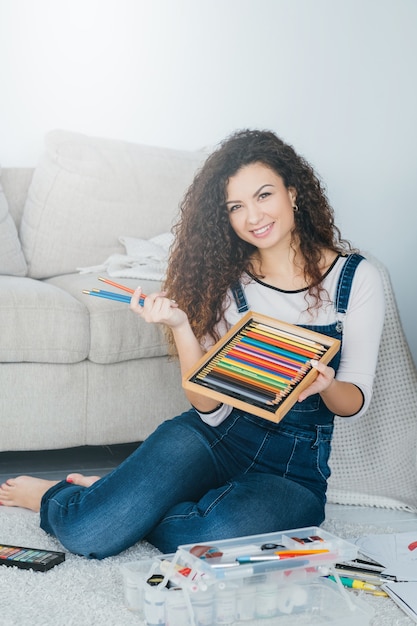 passe-temps d'art à la maison. femme assise sur le sol avec un ensemble de crayons de couleur et un kit de peinture, souriant.