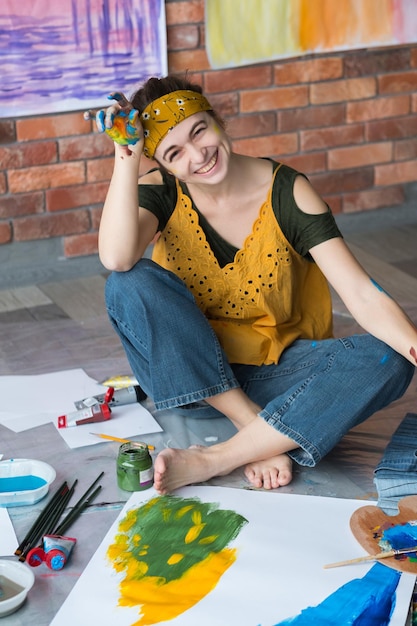 Passe-temps de beaux-arts Jeune artiste féminine créative assise sur le sol les mains sales avec de la peinture en riant