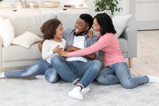 Passe-temps familial. Heureux parents noirs jouant avec leur petite fille à la maison, la chatouillant et s'amusant ensemble dans le salon