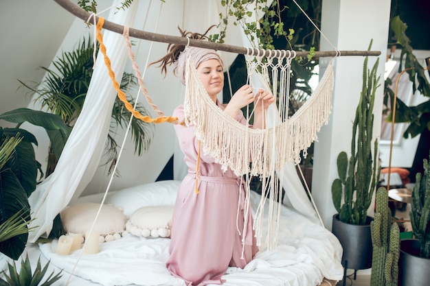 Passe-temps, macramé. Jeune adulte femme affable concentrée en peignoir tissant du macramé sur le lit près des plantes d'intérieur