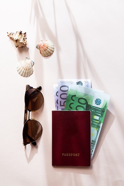 Un passeport avec de l'argent, des lunettes de soleil et des coquillages, à plat sur la table rose clair