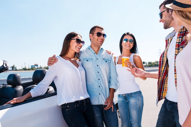 Passer du bon temps avec des amis. Groupe de jeunes gens heureux se parlant et souriant tout en se tenant près de leur cabriolet