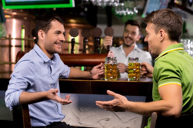 Passer du temps au bar. Deux jeunes hommes buvant de la bière et se parlant pendant que le barman tapotait de la bière en arrière-plan