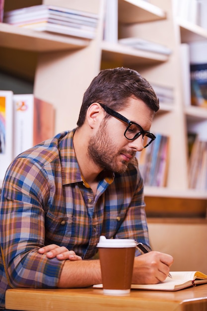 Passer du temps à la bibliothèque. Jeune homme confiant écrivant quelque chose dans le bloc-notes alors qu'il était assis au bureau de la bibliothèque