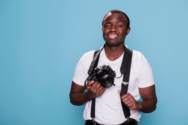 Passionné de photographie d'ethnie africaine joyeuse portant un t-shirt et des bretelles à la mode. Beau photographe professionnel confiant posant en souriant joyeusement et debout sur fond bleu.