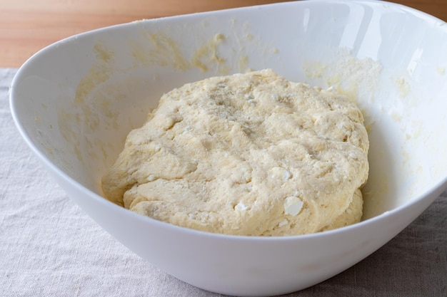 Pâte à caillé crue avec des morceaux de fromage cottage dans un bol blanc sur une nappe en lin sur une table en bois Le processus de fabrication de bagels ou de croissants au caillé