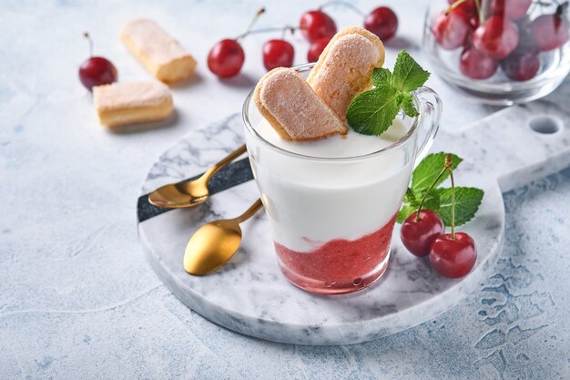 Pâte Feuilletée Aux Cerises D'été Avec Biscuits Savoiardi Et Fromage à La Crème En Verre Sur Fond Gris Clair. Gâteau Tiramisu Traditionnel Avec Des Baies Fraîches. Mise Au Point Sélective.