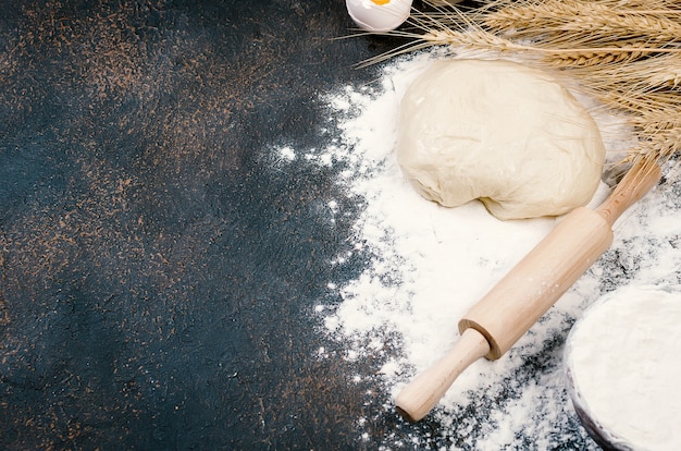 Pâte à levure crue dans un bol et ingrédients, œufs, farine, épis de blé sur une table de cuisine sombre,