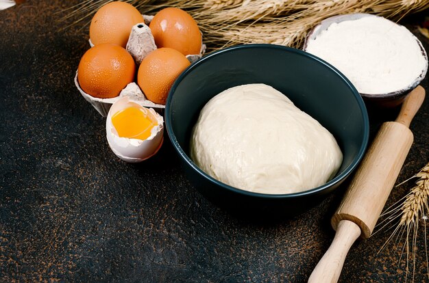 Pâte de levure crue dans un bol et ingrédients, œufs, farine, épis de blé sur table sombre, concept de boulangerie