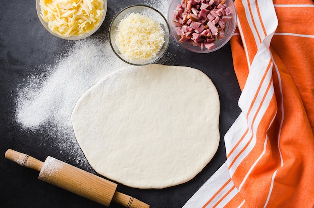 Pâte roulée, farine à pâte, garniture à pizza et rouleau à pâtisserie sur un tableau noir foncé.